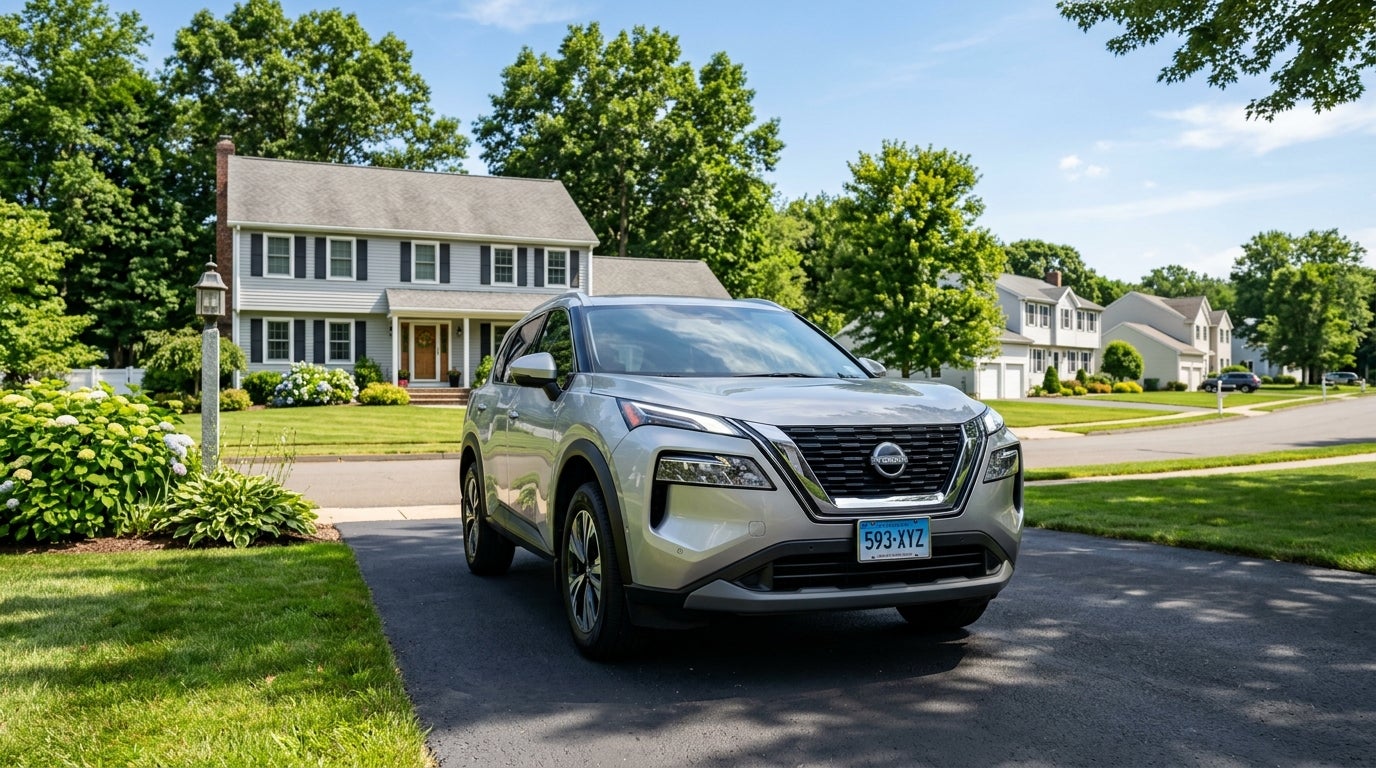 A silver Nissan Rogue parked in the asphalt driveway of a two-story gray house in a suburban neighborhood.