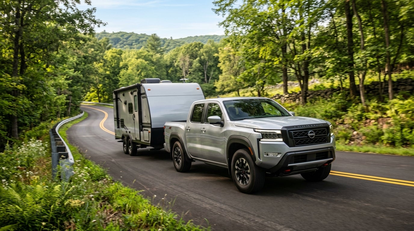Silver Nissan Frontier pickup truck towing a camper trailer on a winding forest road. (Representative image for illustration purposes only. Actual vehicle may vary based on trim and configuration.)