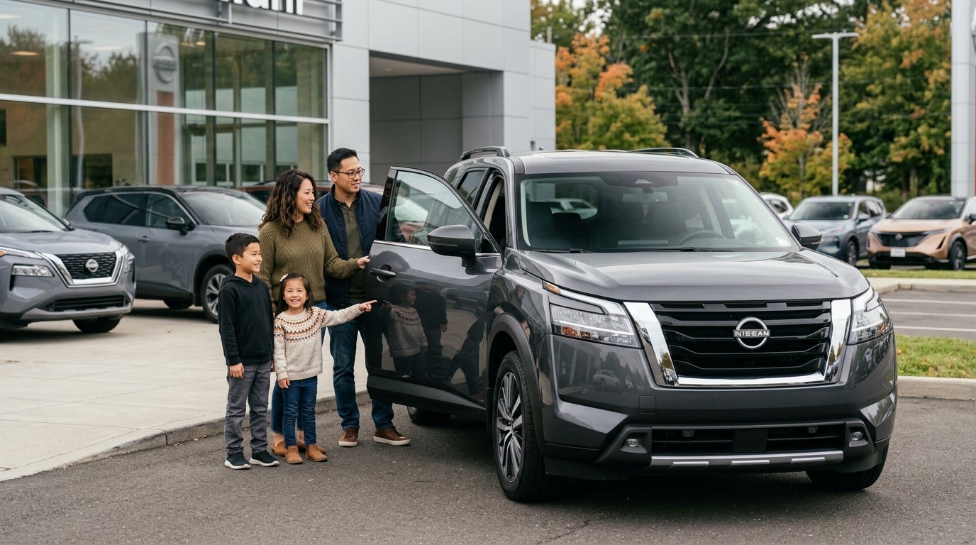 A happy family of four standing next to a dark grey Nissan SUV with an open door at a dealership lot. (Representative image for illustration purposes only. Actual vehicle may vary based on trim and configuration.)